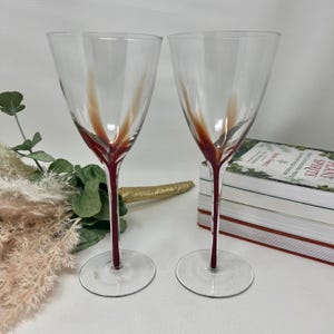 Puede incluir: Dos copas de vino transparentes con un tallo rojo y un diseño rojo que se extiende por el vaso. Las copas están sobre una superficie blanca con una pila de libros y plantas decorativas en el fondo.