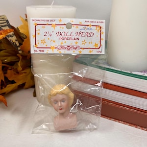 May include: A 5.7 cm porcelain doll head in a clear plastic package. The doll head has blonde hair, closed eyes, and a pink complexion. The package has text that reads "DOLL HEAD PORCELAIN" and "Fibre Craft".