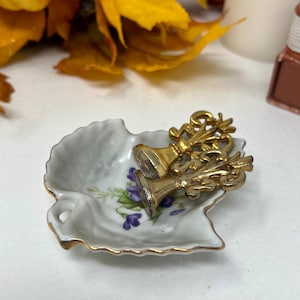 May include: A small, leaf-shaped dish with a white base, gold trim, and a floral design featuring purple flowers. Two gold-colored decorative objects rest inside the dish. The background includes yellow leaves and a white surface.