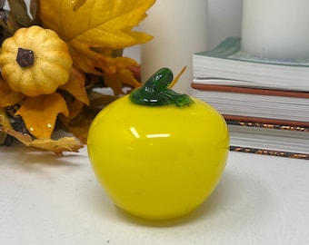 May include: A bright yellow glass apple with a green stem and leaf. The apple is sitting on a white surface.