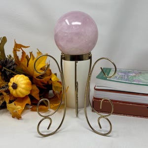 Può includere: Una sfera di quarzo rosa, di circa 10 cm di diametro, esposta su un supporto decorativo in metallo color ottone. Il supporto ha un design curvo e aperto. La sfera è di colore rosa tenue e poggia su una base cilindrica color crema.