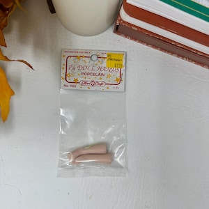 May include: A package of porcelain doll hands. The package has the text "F/8 DOLL HANDS PORCELAIN" and "Fibre Craft" printed on it. The doll hands are a light pink color. The package is clear plastic.