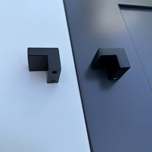 Puede incluir: Dos tiradores de puerta de armario de metal negro con forma cuadrada y un ángulo de 90 grados. Los tiradores están montados en una superficie blanca y azul.