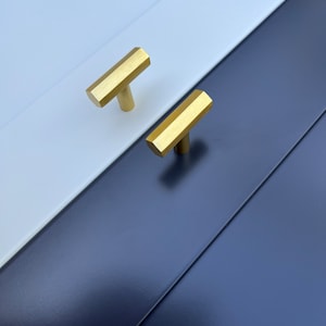 May include: Two gold cabinet knobs with a hexagonal shape and a T-bar design. The knobs are mounted on a white and navy blue cabinet door.