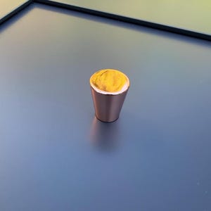 Pode incluir: Um objeto em forma de cone de cor cobre com um topo amarelo texturizado e uma borda branca. O objeto está sobre uma superfície azul-cinza, com uma borda escura no fundo. O objeto parece ser um elemento decorativo.