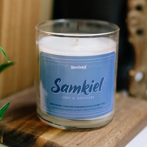 May include: A clear glass candle jar with a blue label that reads "Samkiel Gods & Monsters". The candle sits on a small wooden cutting board. The label also lists the scent as "Chocolate | Toasted Marshmallow | Divine Fury".