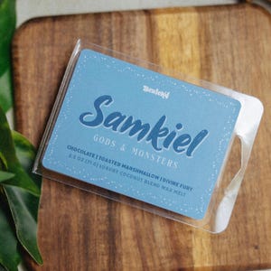 May include: A blue wax melt package with the name "Samkiel" and the words "Gods & Monsters" printed on it. The package contains a 2.5 oz (71 g) luxury coconut blend wax melt with chocolate and toasted marshmallow scents.