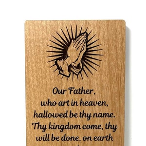 May include: A wooden bookmark with a laser-engraved image of praying hands and the Lord's Prayer. The text reads: "Our Father, who art in heaven, hallowed be thy name. Thy kingdom come, thy will be done, on earth as it is in heaven. Give us this day our daily bread. And forgive us our trespasses, as we forgive those who trespass against us. And lead us not into temptation, but deliver us from evil. For thine is the kingdom, and the power, and the glory, for ever and ever. Amen."