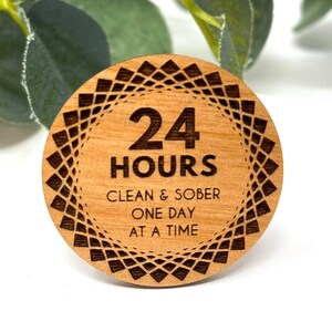 Puede incluir: Un token redondo de madera con un dise&ntilde;o geom&eacute;trico en blanco y negro alrededor del borde. El centro del token tiene el texto "24 HOURS" en letras negras, seguido de "CLEAN & SOBER ONE DAY AT A TIME" en letras negras m&aacute;s peque&ntilde;as.
