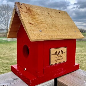 Puede incluir: Una casita de p&aacute;jaros de madera roja con un techo de madera marr&oacute;n. La casita tiene un peque&ntilde;o agujero para que los p&aacute;jaros puedan entrar y un letrero que dice "every little thing is gonna be alright".