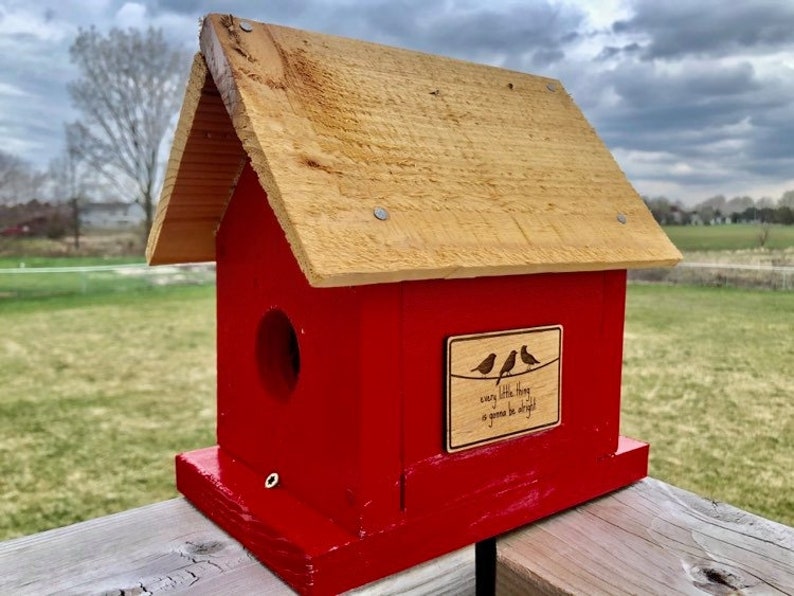 Puede incluir: Una casita de p&aacute;jaros de madera roja con un techo de madera marr&oacute;n. La casita tiene un peque&ntilde;o agujero para que los p&aacute;jaros puedan entrar y un letrero que dice "every little thing is gonna be alright".