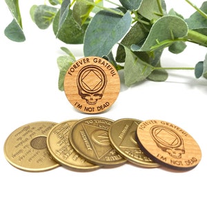 May include: Five wooden and metal tokens. Four of the tokens are gold colored and have text and a symbol. One token is made of wood and has the text "FOREVER GRATEFUL I'M NOT DEAD" and a skull and crossbones symbol.