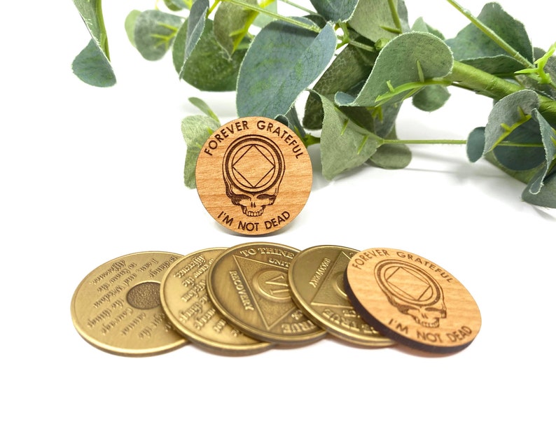May include: Five wooden and metal tokens. Four of the tokens are gold colored and have text and a symbol. One token is made of wood and has the text "FOREVER GRATEFUL I'M NOT DEAD" and a skull and crossbones symbol.