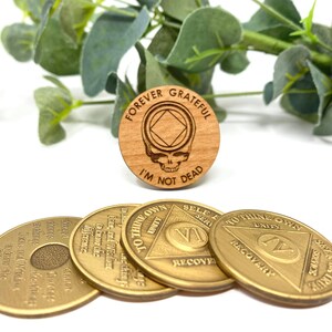 May include: Four gold-colored metal coins with the words "To Thine Own Self Be True" and "Unity" on them. The coins are arranged in a circle with a wooden coin in the center. The wooden coin has a skull and crossbones design with the words "Forever Grateful I'm Not Dead" on it.