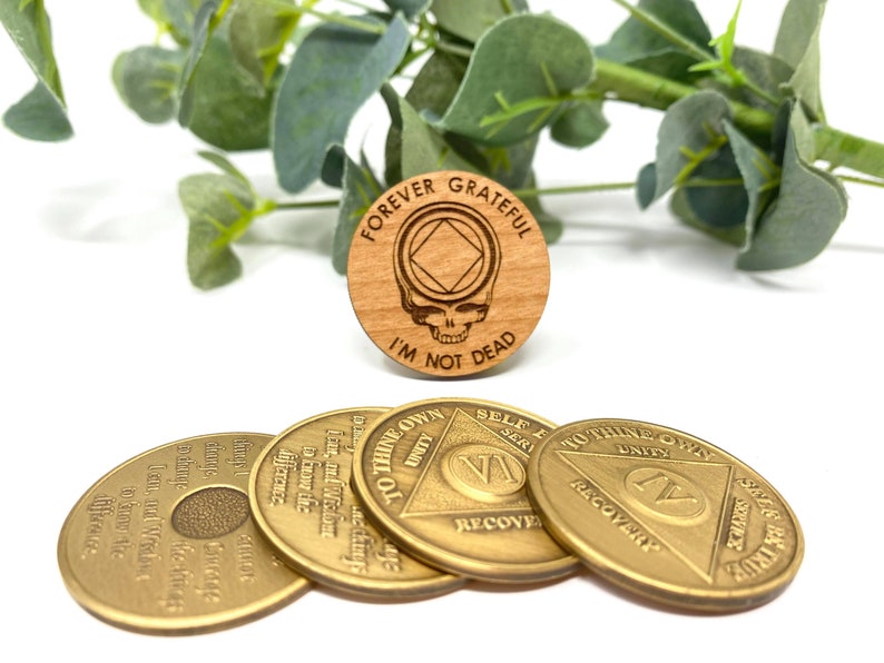 May include: Four gold-colored metal coins with the words "To Thine Own Self Be True" and "Unity" on them. The coins are arranged in a circle with a wooden coin in the center. The wooden coin has a skull and crossbones design with the words "Forever Grateful I'm Not Dead" on it.