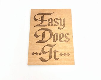 Puede incluir: Un letrero de madera con el texto "Easy Does It" en una fuente decorativa. El letrero tiene una veta de madera marrón claro y el texto es marrón oscuro.