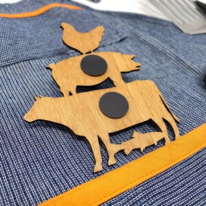 May include: Wooden cutout of a cow, pig, and chicken stacked on top of each other. The cutout is attached to a blue and white denim apron with an orange stripe. Two black magnets are attached to the back of the cutout.