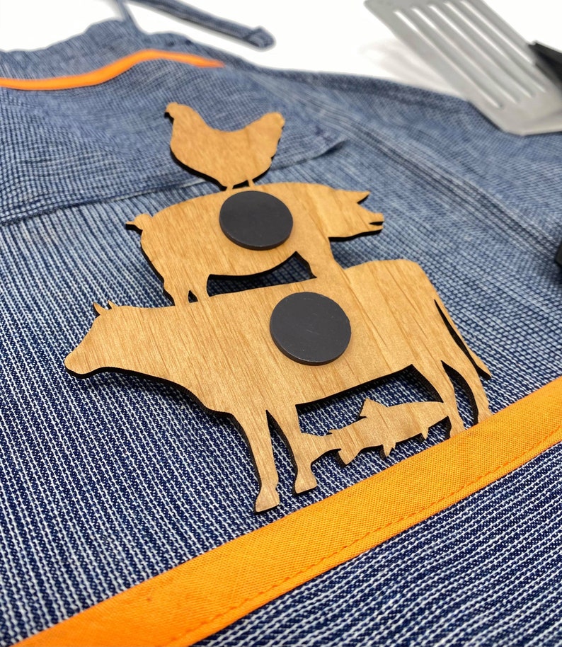 May include: Wooden cutout of a cow, pig, and chicken stacked on top of each other. The cutout is attached to a blue and white denim apron with an orange stripe. Two black magnets are attached to the back of the cutout.