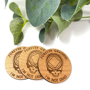 May include: Three wooden round coasters with a Grateful Dead inspired design. Each coaster features a skull with a diamond shape in the forehead and the text "Forever Grateful I'm Not Dead".