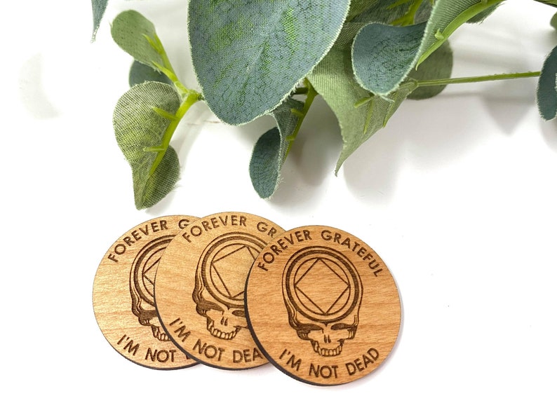 May include: Three wooden round coasters with a Grateful Dead inspired design. Each coaster features a skull with a diamond shape in the forehead and the text "Forever Grateful I'm Not Dead".