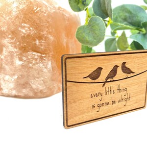 Puede incluir: Un letrero de madera con veta de madera marr&oacute;n claro y borde marr&oacute;n oscuro. Tres p&aacute;jaros est&aacute;n posados en un alambre con el texto "every little thing is gonna be alright".