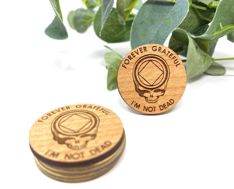May include: Two wooden round coasters with the text "Forever Grateful I'm Not Dead" and a skull with a diamond shape in the middle. The coasters are made of light brown wood.