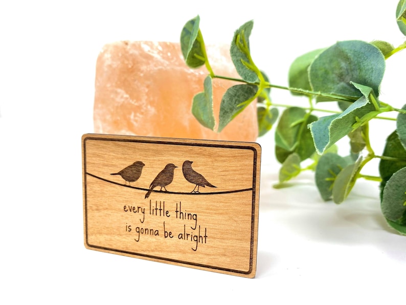 Puede incluir: Un letrero de madera con un acabado de madera de grano marr&oacute;n claro. El letrero presenta tres p&aacute;jaros posados ​​en un alambre con el texto "every little thing is gonna be alright".