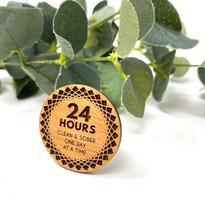 Puede incluir: Un token redondo de madera con un dise&ntilde;o geom&eacute;trico alrededor del borde. El centro del token dice "24 HORAS LIMPIO Y SOBRIO UN D&Iacute;A A LA VEZ".
