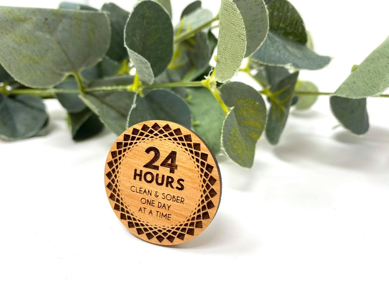 Puede incluir: Un token redondo de madera con un dise&ntilde;o geom&eacute;trico alrededor del borde. El centro del token dice "24 HORAS LIMPIO Y SOBRIO UN D&Iacute;A A LA VEZ".