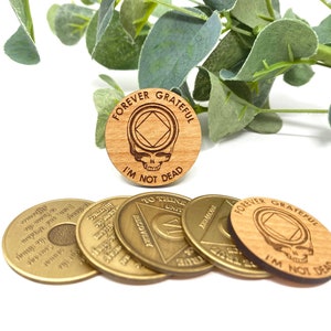 May include: Five wooden tokens with the text "Forever Grateful I'm Not Dead" and a skull and crossbones graphic. The tokens are brown and have a wood grain texture. Four of the tokens are gold colored and have text and a triangle graphic.