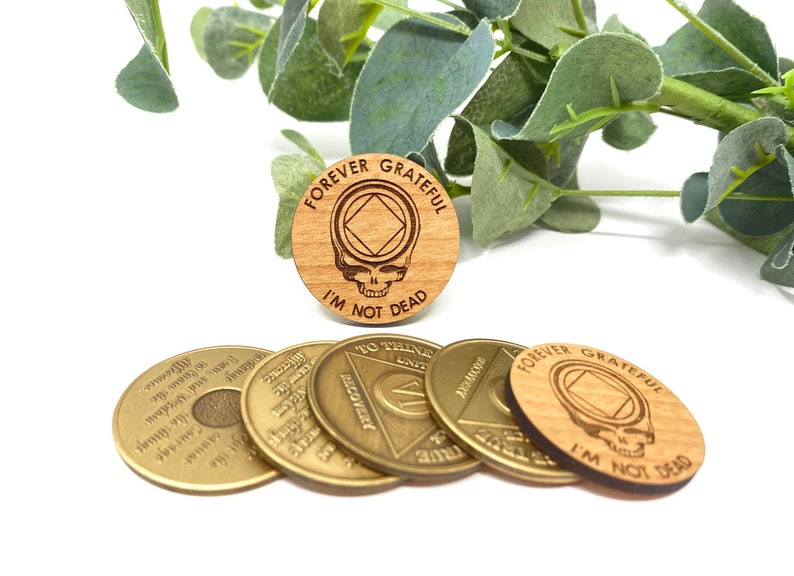 May include: Five wooden tokens with the text "Forever Grateful I'm Not Dead" and a skull and crossbones graphic. The tokens are brown and have a wood grain texture. Four of the tokens are gold colored and have text and a triangle graphic.