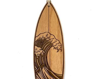 Op de afbeelding: Een houten surfplank ornament met een gedetailleerde carving van een grote brekende golf. Het ornament is bruin en heeft een touwtje om op te hangen.