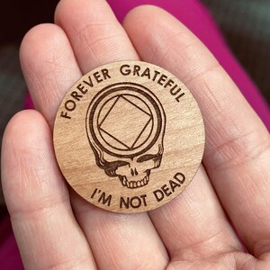May include: A wooden token with a Grateful Dead skull and the text "Forever Grateful I'm Not Dead" and a square inside a circle.
