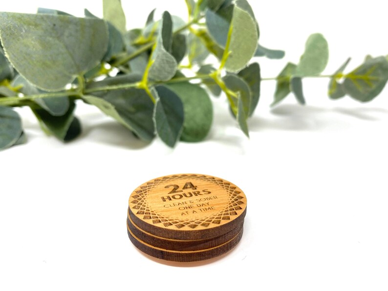 Puede incluir: Un posavasos redondo de madera con el texto "24 HOURS CLEAN & SOBER ONE DAY AT A TIME" grabado en &eacute;l. El posavasos tiene un patr&oacute;n decorativo alrededor del borde.