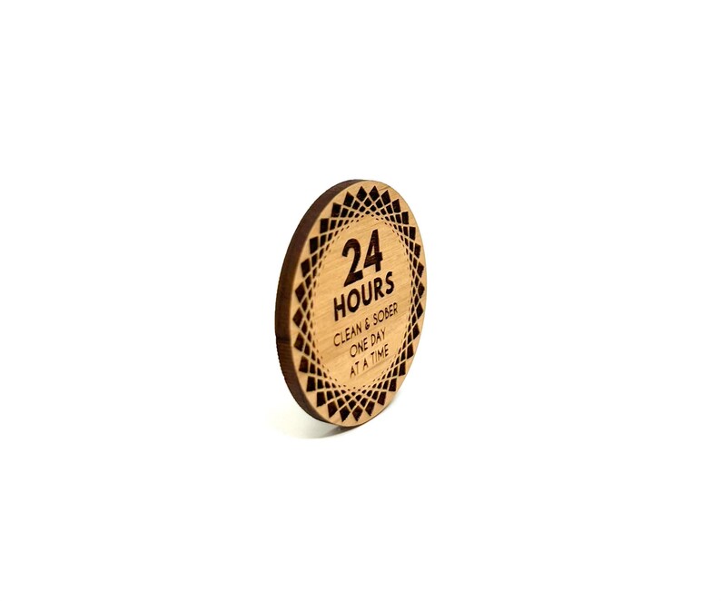 Puede incluir: Un token redondo de madera con un dise&ntilde;o geom&eacute;trico en blanco y negro. El token tiene el texto "24 HOURS" y "CLEAN & SOBER ONE DAY AT A TIME" grabado en &eacute;l.