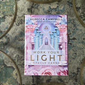 Op de afbeelding: Een rechthoekig kaartspel Work Your Light orakelkaarten van Rebecca Campbell met illustraties van Danielle Noel. De doos toont een roze en blauwe afbeelding van een stenen tempelpad, een gloeiend driehoekssymbool en grote roze rozen.