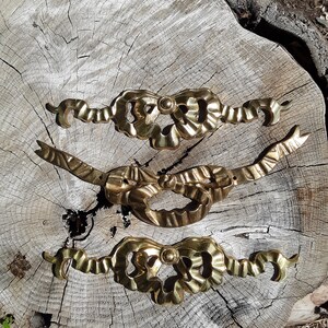 Puede incluir: Tres piezas decorativas de metal dorado en forma de lazos con detalles intrincados. Las piezas están dispuestas en fila sobre una superficie de madera.