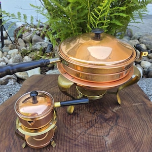 May include: A large copper chafing dish with a lid, a long black handle, and a brass stand, accompanied by a smaller matching copper pot with a black handle and lid on a brass base. Both pieces feature polished copper surfaces and black knobs.