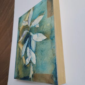 K&ouml;nnte beinhalten: Abstrakte Malerei auf einem Holzrahmen. Das Kunstwerk zeigt ein zentrales wei&szlig;es Blumendesign vor einem Hintergrund aus T&uuml;rkis-, Blau- und Braunt&ouml;nen. Die R&auml;nder des Rahmens sind naturfarbenes Holz.