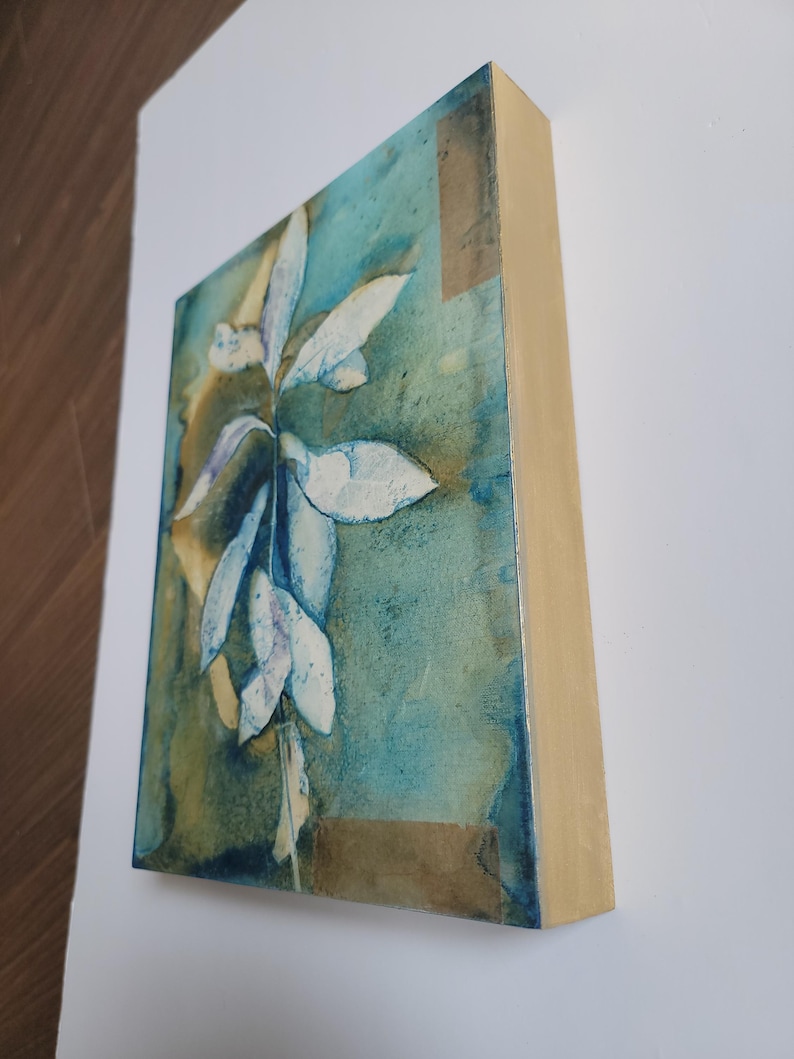K&ouml;nnte beinhalten: Abstrakte Malerei auf einem Holzrahmen. Das Kunstwerk zeigt ein zentrales wei&szlig;es Blumendesign vor einem Hintergrund aus T&uuml;rkis-, Blau- und Braunt&ouml;nen. Die R&auml;nder des Rahmens sind naturfarbenes Holz.