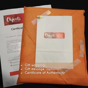 K&ouml;nnte beinhalten: Ein orangefarbenes Geschenkpaket mit einer wei&szlig;en Karte und einem Echtheitszertifikat. Das Paket ist mit dekorativem Klebeband versiegelt. Die wei&szlig;e Karte tr&auml;gt den Text "Objects Art Gallery" und ein Zertifikat.