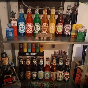 Peut inclure: Une étagère avec une variété de bouteilles en verre colorées, certaines avec des étiquettes qui disent "Juggernog", "Speed Cola", "Double Tap", "Mule Kick" et "Ph.D. Flopper". Il y a aussi plusieurs petites bouteilles sur l'étagère inférieure, et une boîte de jeu Call of Duty.