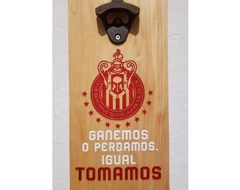 May include: A wooden bottle opener with a red and white logo of a soccer team. The logo says "Club Deportivo Guadalajara" and has a crest with a crown and a lion. The text below the logo says "Ganemos o Perdamos, Igual Tomamos".