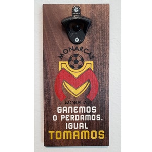May include: Wooden bottle opener with a black metal opener and a logo for the soccer team Monarcas Morelia. The logo features a red and yellow "M" with a soccer ball inside. The text below the logo reads "Ganemos o perdamos, igual tomamos".