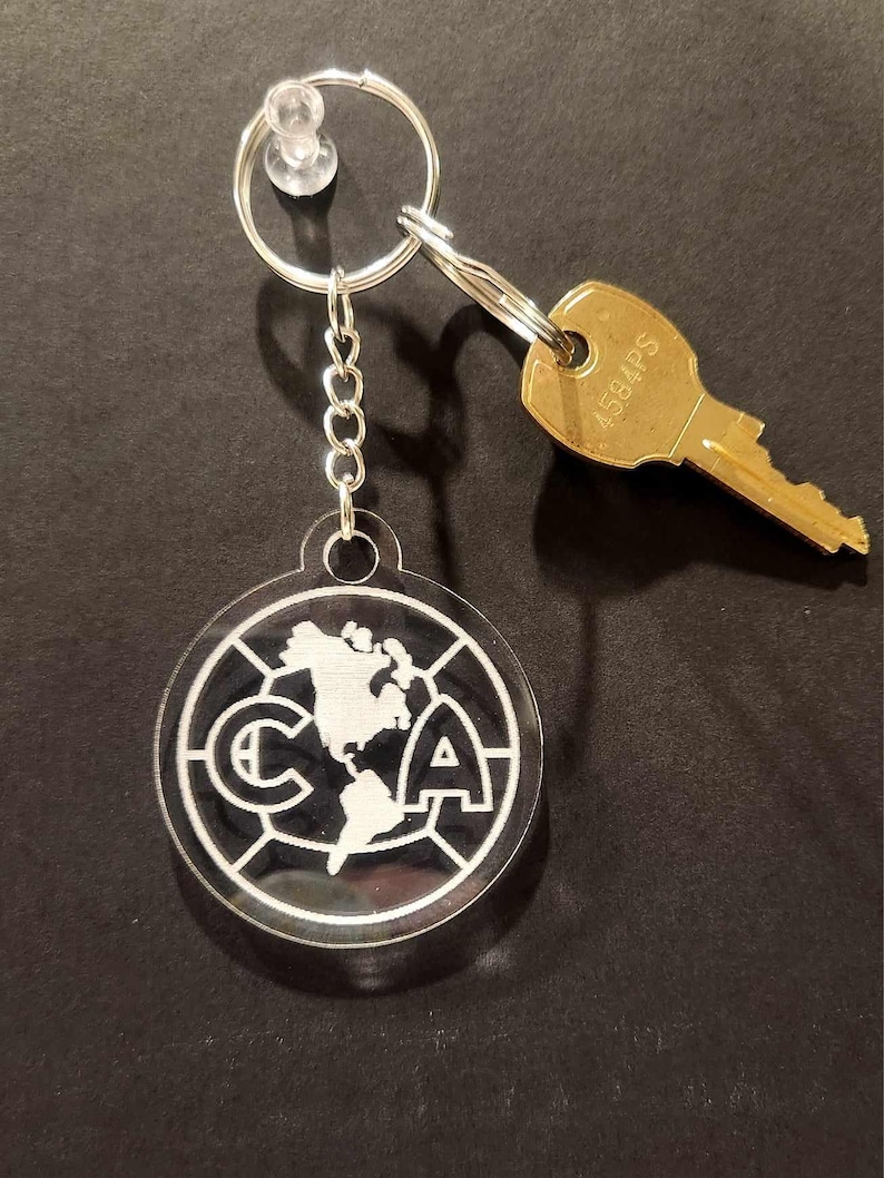 Pode incluir: Um chaveiro prateado com um charme circular de acr&iacute;lico preto e branco que apresenta o logotipo do Club Am&eacute;rica, um time de futebol profissional mexicano. O logotipo inclui as letras "CA" e uma imagem estilizada de uma bola de futebol com um mapa do mundo dentro.