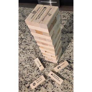 May include: A wooden block stacking game with engraved text. The tower is partially built, with several blocks scattered around. Text on the blocks includes phrases like "Everyone Drinks!" and "Skip your next turn!" A fun party game.