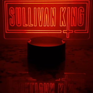 May include: A red neon sign with the text "Sullivan King" in a rectangular frame. The sign is lit up and is sitting on a black base.
