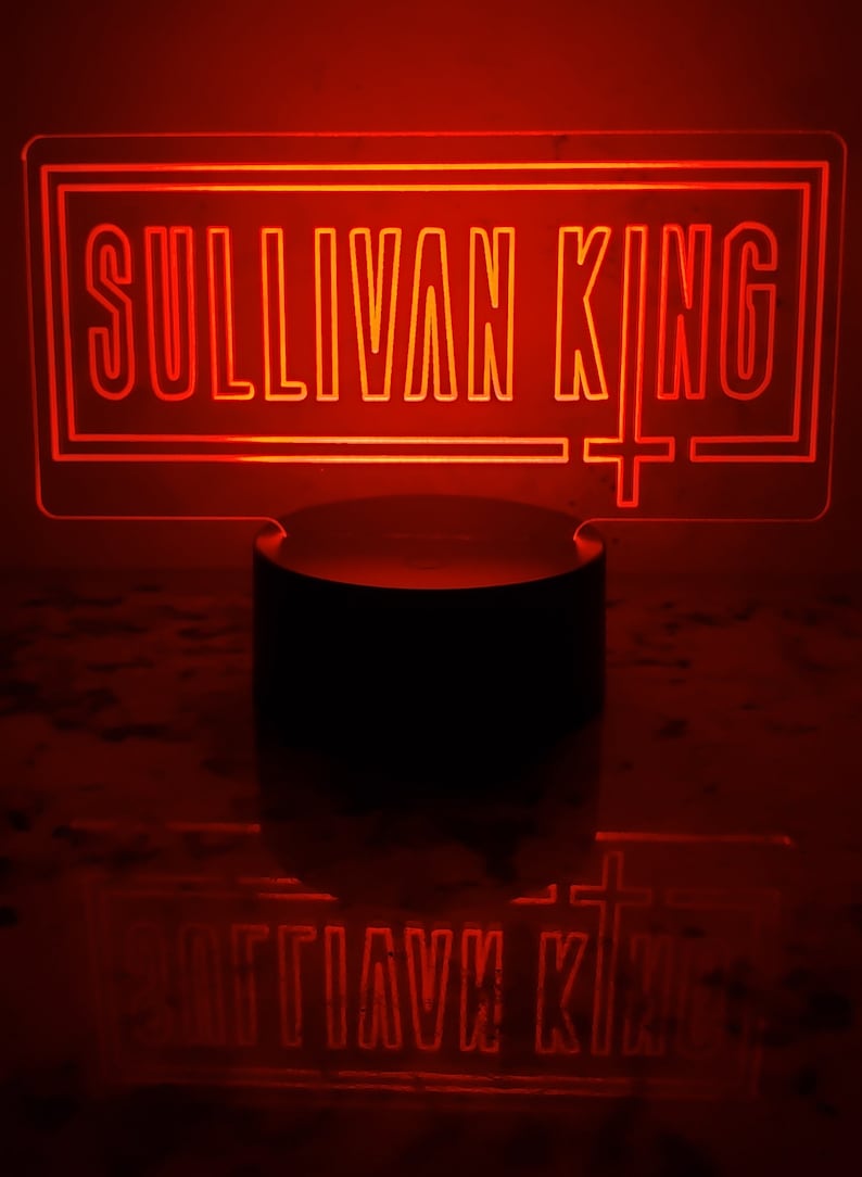 May include: A red neon sign with the text "Sullivan King" in a rectangular frame. The sign is lit up and is sitting on a black base.