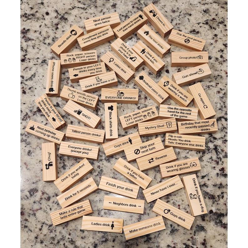 May include: A collection of wooden blocks with various drinking game instructions printed on them. The blocks are scattered on a speckled surface. Text includes phrases like "Drink if you have a dog" and "Everyone drinks!"