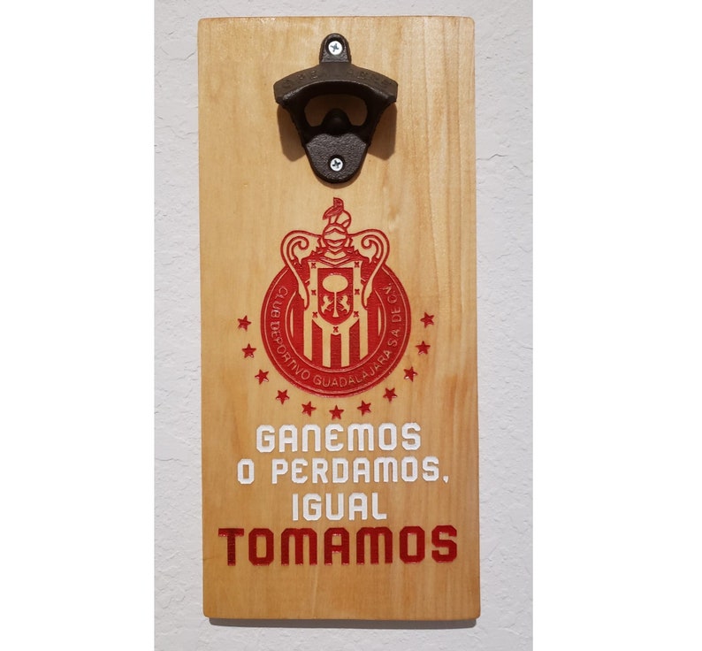 Puede incluir: Un abridor de botellas de madera con un logotipo rojo y blanco de un equipo de f&uacute;tbol. El logotipo dice "Club Deportivo Guadalajara" y tiene un escudo con una corona y un le&oacute;n. El texto debajo del logotipo dice "Ganemos o Perdamos, Igual Tomamos".